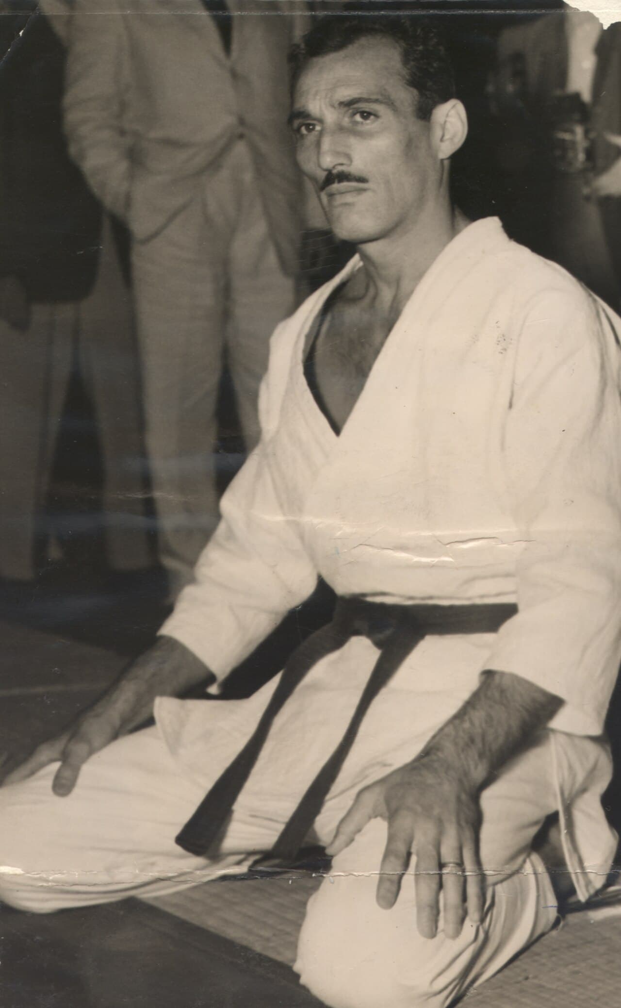 Black and white photograph of Helio Gracie kneeling on a mat in a white gi and dark belt, catalogued in 1952.
