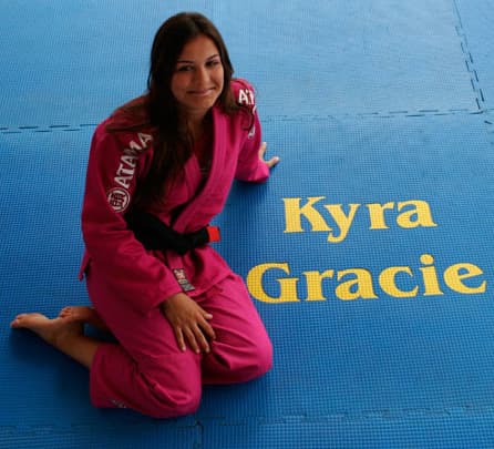 Kyra Gracie kneeling on a blue mat in a pink gi and black belt, with her name lettered on the mat beside her.