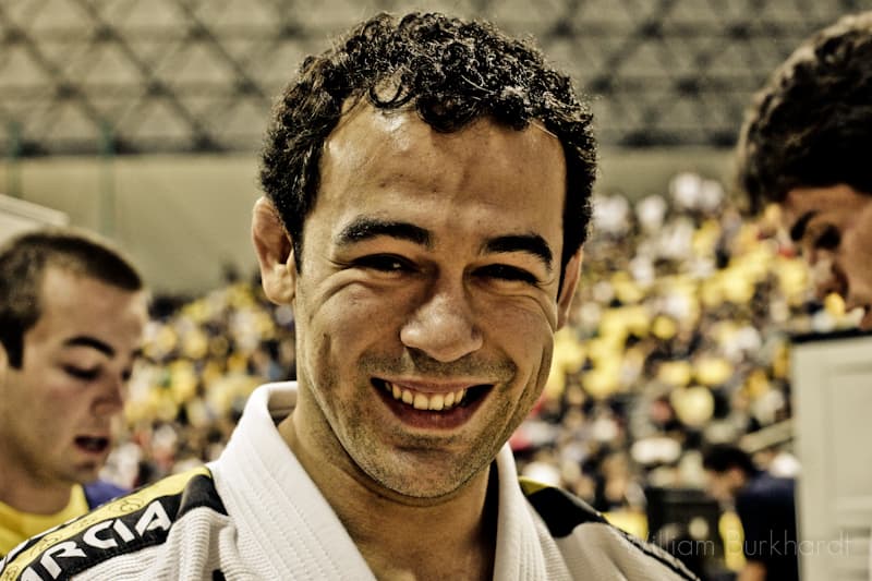 Marcelo Garcia photographed in a white gi at a crowded championship venue, smiling towards the camera.