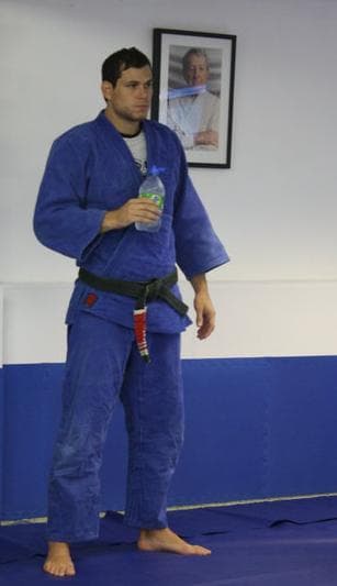 Roger Gracie standing barefoot on the mats in a blue gi and black belt during a class at a gym in Woking, England.