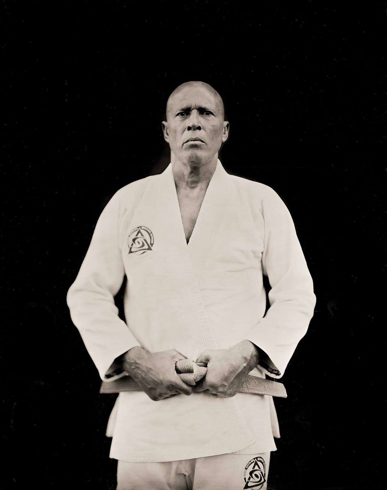 Wet plate collodion portrait of Royce Gracie standing in a white gi against a black background, hands folded at his belt.